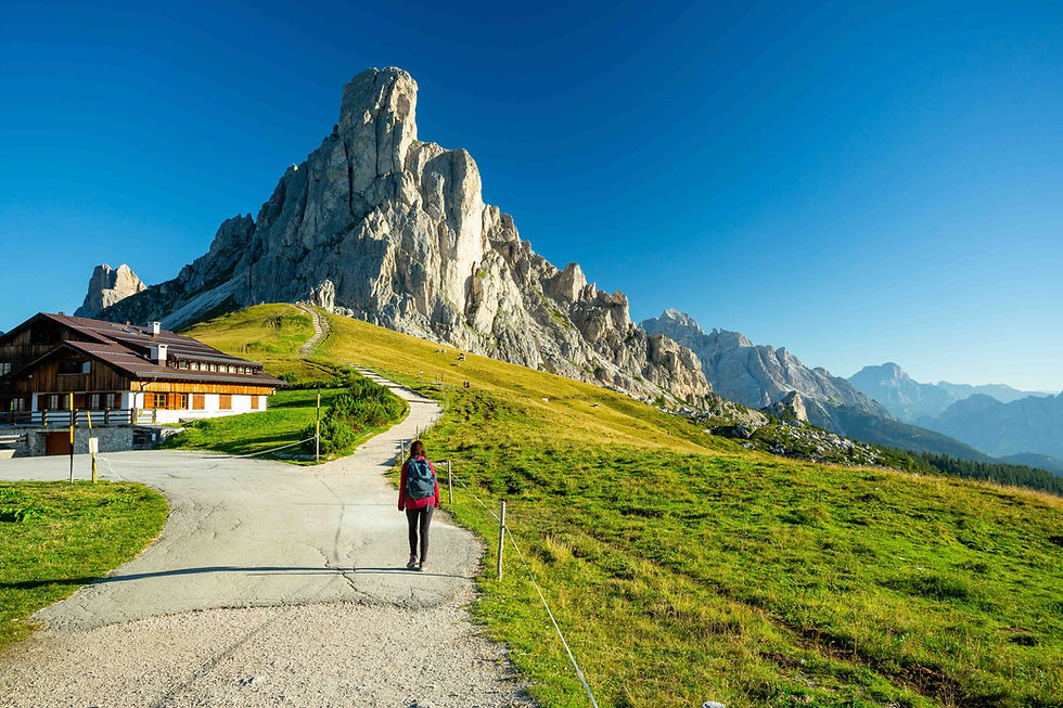 Hiker walks up a winding path past a chalet toward a towering rocky mountain under a clear blue sky.