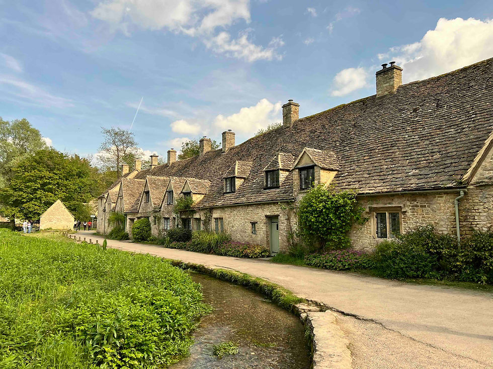Stone cottages with steep roofs beside a winding stream and path under a blue sky with clouds; peaceful countryside scene