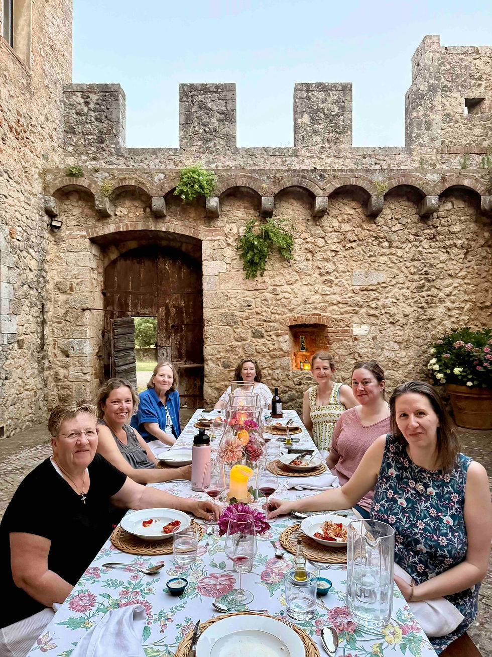 Seven women share a candlelit meal at a floral table in a stone courtyard of an old fortress, smiling at the camera.