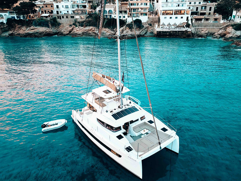 White catamaran with a small dinghy floats in a turquoise cove, with cliffside seaside buildings in the background.