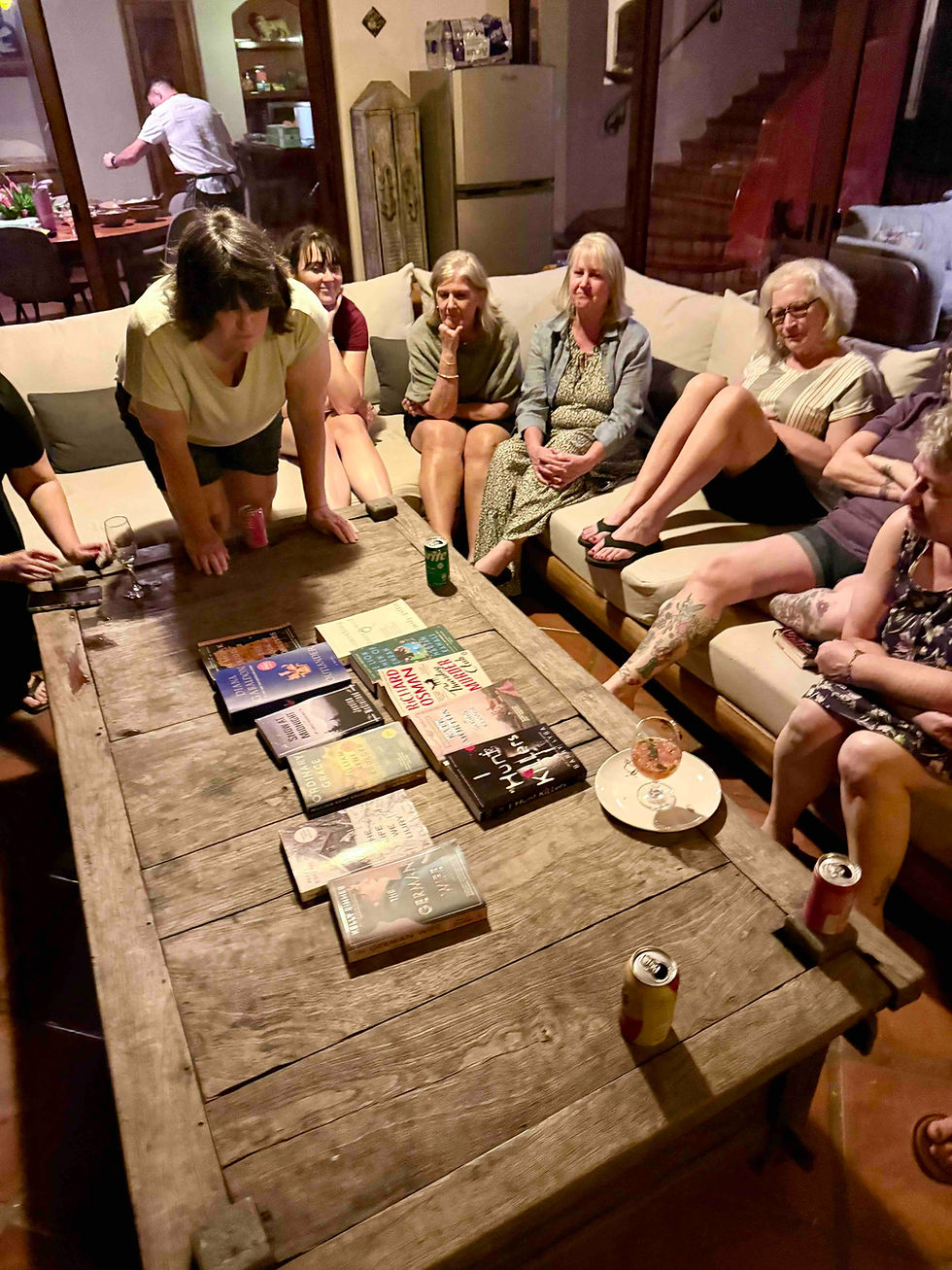 Women gather on a sofa around a wooden table with books and drinks. A man cooks in the background. The room feels relaxed and engaging.