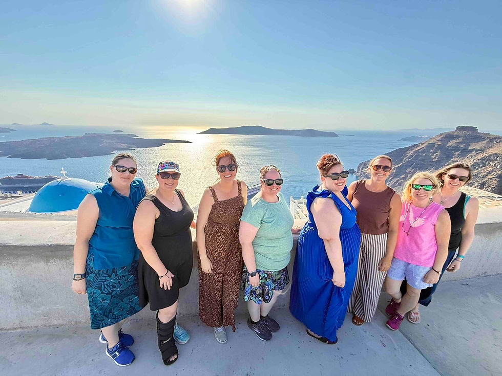 Seven smiling women in sunglasses pose on a sunny cliffside terrace overlooking a blue sea and rocky islands.