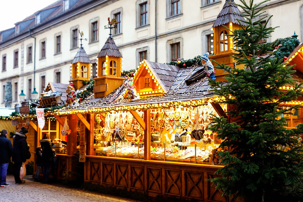 Lit Christmas market stall with gingerbread hearts and pastries, shoppers browsing, beside a pale stone building and pine tree.