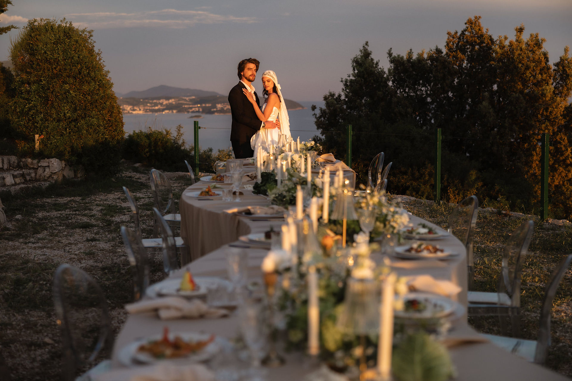 A destination wedding couple standing together at the end of a long luxury wedding reception table at dusk, with candlelights glowing and the sea in the background at Park Orsula, Dubrovnik.