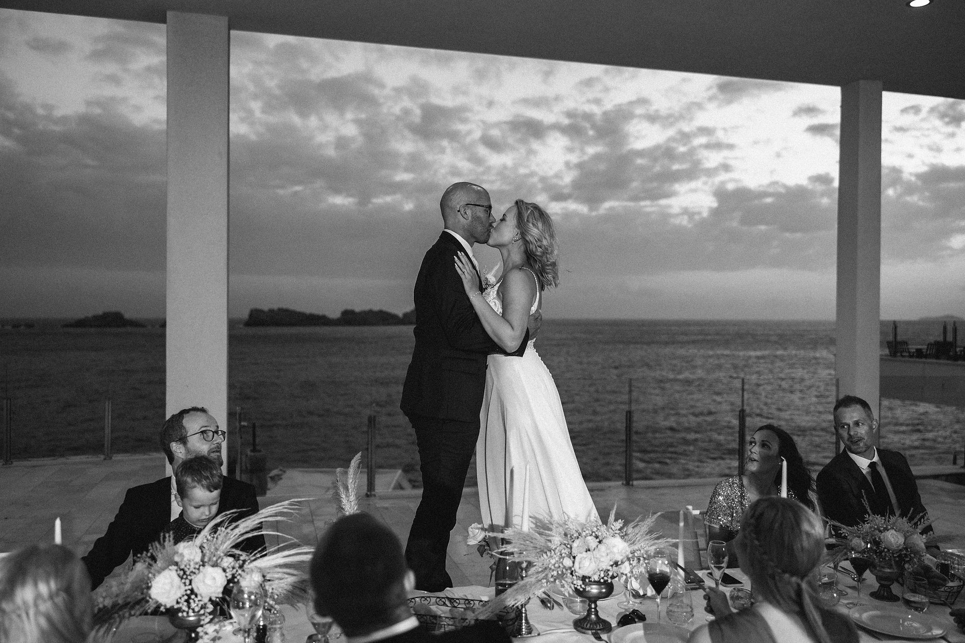 bride and groom standing on chairs during dinner on the terrace of a restaurant by the sea