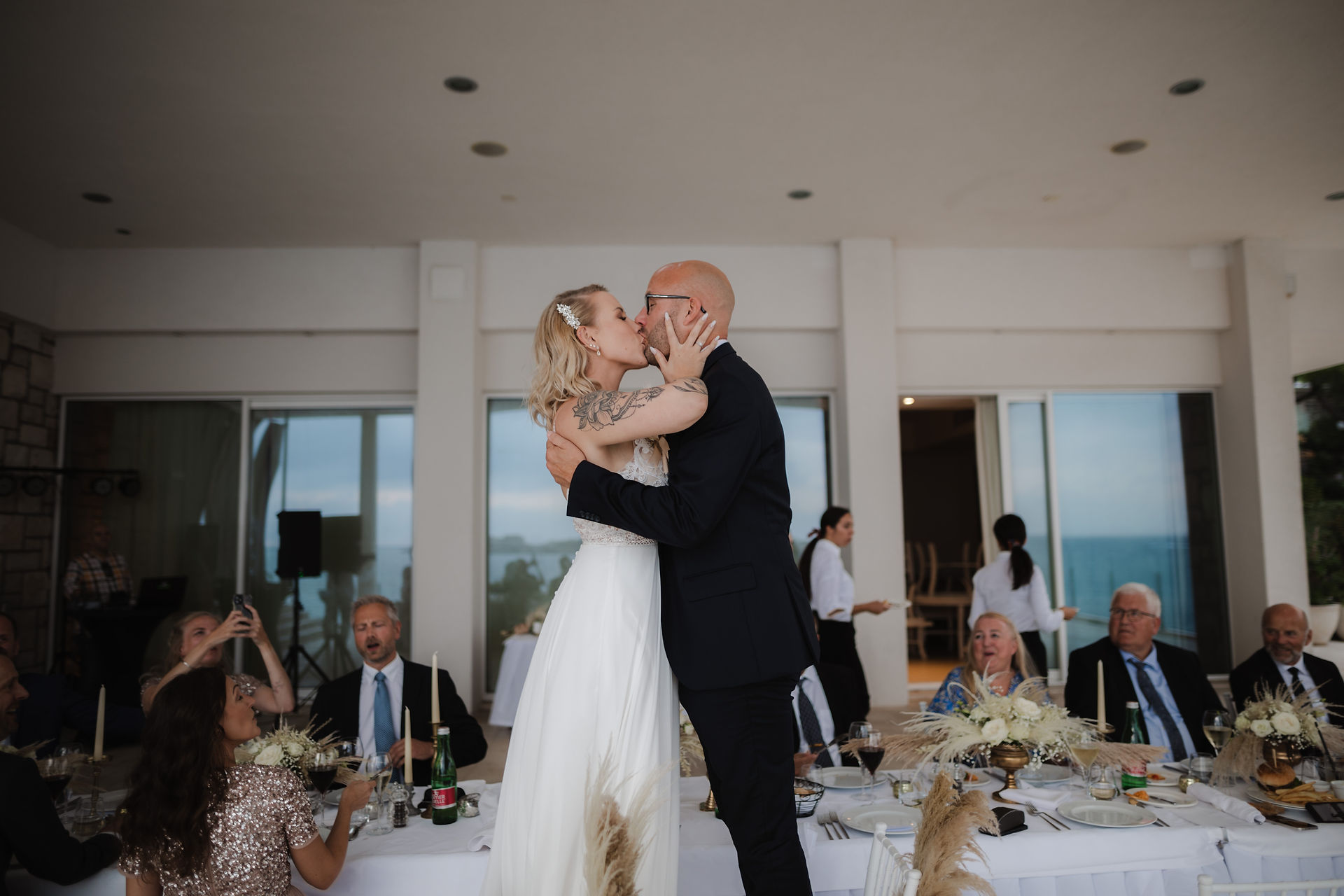 bride and groom stand on chairs and kiss during dinner on the terrace of a restaurant by the sea