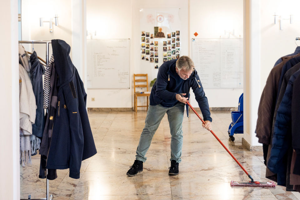 Ein Mann wischt mit einem Bodenwischer den Marmorboden in einem hellen Flur mit Garderobenständern und Whiteboards.
