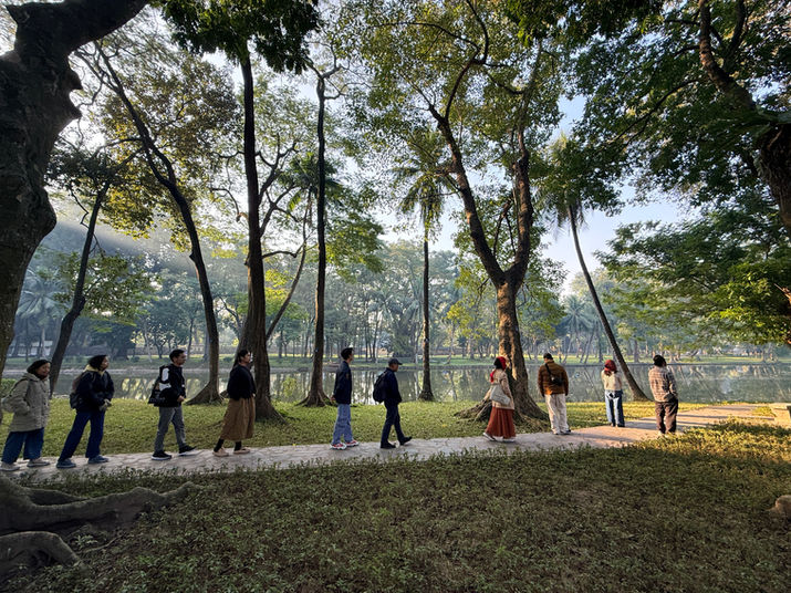 People stroll single file through a picturesque park, surrounded by tall, green trees and a tranquil lake in the background. The clear morning air and sunlight filtering through the canopy create a peaceful atmosphere.