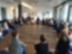 A large group of people sit in a wide circle on meditation cushions in a bright seminar room. Everyone is looking attentively towards the center, some taking notes. The room feels calm, focused, and inviting – like a mindfulness or dialogue circle.