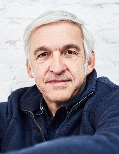 Ein Mann mit weißem Haar und leichtem Lächeln schaut ruhig in die Kamera. Er trägt einen dunkelblauen Pullover und sitzt vor einer weißen Backsteinwand.