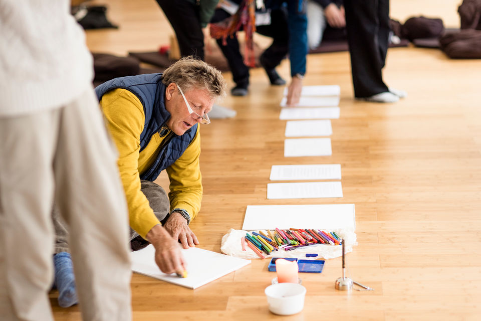 Ein Mann kniet auf einem Holzboden und zeichnet, vor ihm liegen bunte Stifte, Papierblätter und eine Kerze.“