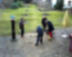 Achtsame Arbeit Fünf Personen arbeiten gemeinsam im Freien auf einem gepflasterten Platz vor einer Wiese. Eine Person schiebt eine Schubkarre, zwei kehren den Boden, eine hält ein Werkzeug mit langem Stiel, und eine weitere Person beobachtet die Gruppe. Alle tragen wetterfeste Kleidung. Im Hintergrund sind eine Wasserpumpe, ein geschlossener Sonnenschirm und ein Garten mit kleinen Bäumen zu sehen.