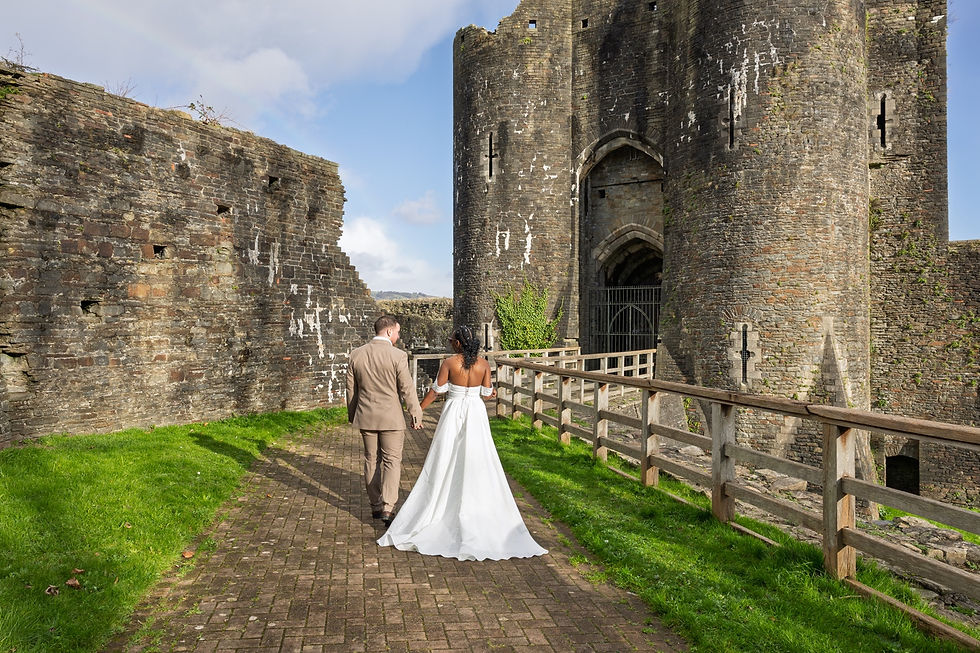 Bride and groom walking hand in hand toward a stone castle gate on a brick path, with green grass and blue sky.