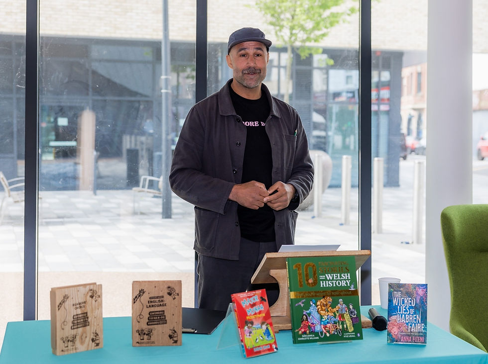 Man standing indoors by a table with various books displayed. Background shows large windows overlooking a street. Bright and casual setting.