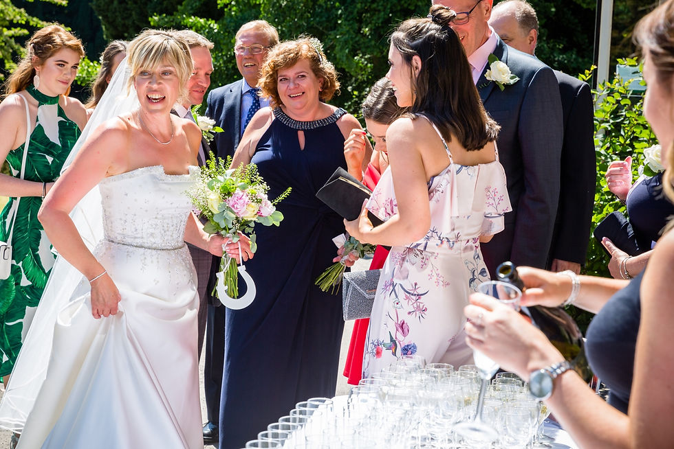 Bride in white dress holds flowers and horseshoe, surrounded by smiling guests outdoors. Woman pours champagne into glasses. Sunny setting.