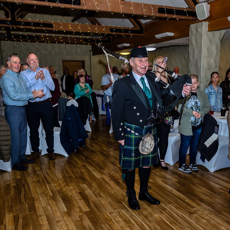 Bagpiper in tartan kilt performs at an indoor celebration as guests stand and applaud under string lights.