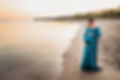 A pregnant woman in a green dress stands on a beach at waters edge at sunrise
