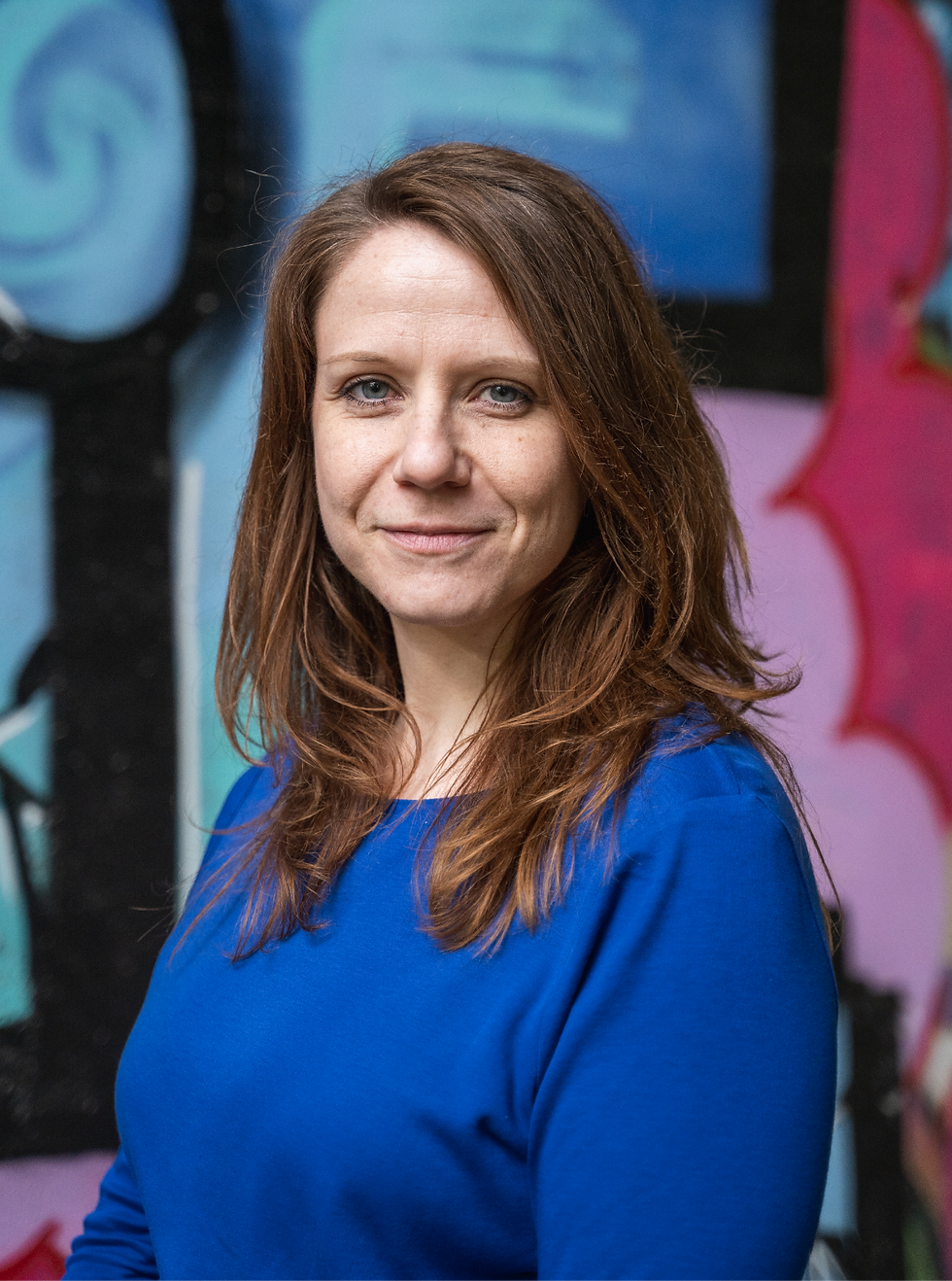 Bild der Autorin Doris Moravec. Sie hat schulterlange braune Haare, die sie offen trägt und sie schaut lächelnd in die Kamera. Sie trägt ein blaues, langärmliges Oberteil. Sie steht vor einer Wand mit blau/pinken Graffiti