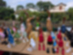 Colorful wooden duck figurines in clothes on a table at an outdoor market, with houses and trees in the background.