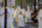 a young girl in white and cream religious robing marches along with a procession of cream hooded men