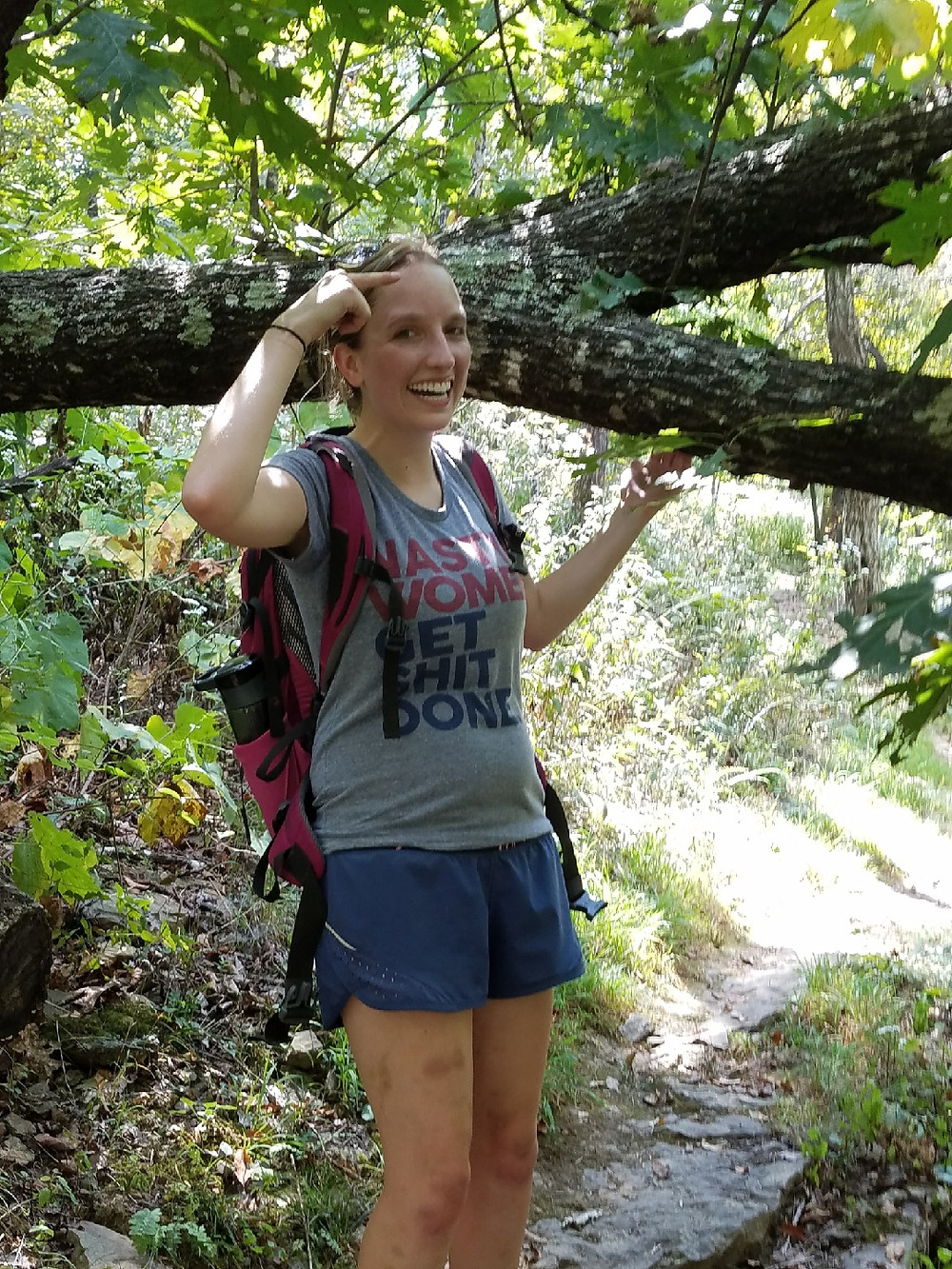 Shannon poses in front of a large tree branch next to her. She is smiling and pointing at her head. She is dressed in hiking clothing - a t--shirt, shorts and a camelback backpack.