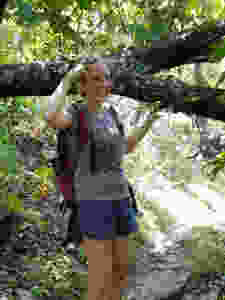 Shannon poses in front of a large tree branch next to her. She is smiling and pointing at her head. She is dressed in hiking clothing - a t--shirt, shorts and a camelback backpack.