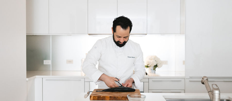 Chef Victor Rivera fillets a fish on a wooden board in a bright white kitchen, focused and calm.