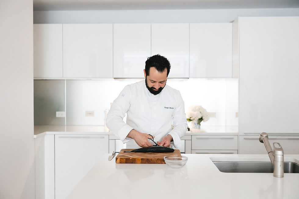 Chef Victor Rivera fillets a fish on a wooden board in a bright white kitchen, focused and calm.