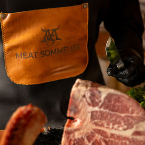 Gloved meat sommelier in black apron with MEAT SOMMELIER text holds a knife beside raw marbled beef in a warm kitchen setting