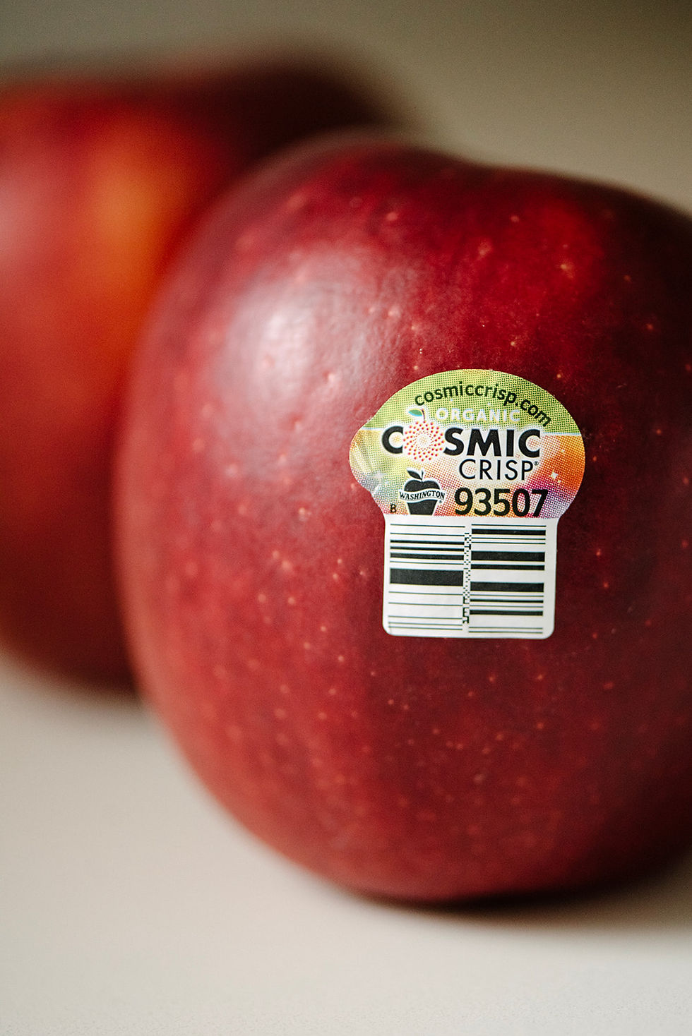 Close-up of two red apples, one with a sticker reading "Cosmic Crisp 93507." The background is soft and neutral.
