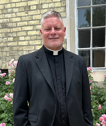 Rev. Don Beyers stands outside Grace Church on-the-Hill in front of a bed of pink roses, and smiles at the camera. He's dressed in black.
