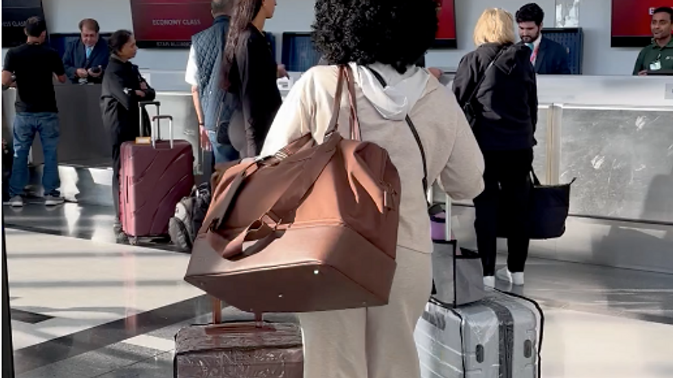 Traveler in beige outfit at airport check-in, carrying brown Beis weekender bag and two Beis luggage.