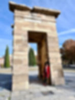 A black woman in a red Zara coat leans against Temple of Debod in Madrid, Spain. Clear blue sky, trees, and distant buildings create a tranquil backdrop.