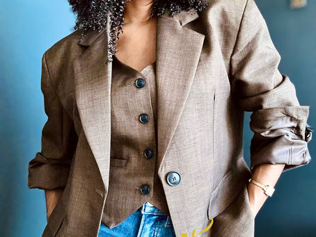 Woman with curly hair, red lipstick, wearing a brown blazer and matching vest over jeans, stands against a dark blue wall. Text: Voyage à nue.