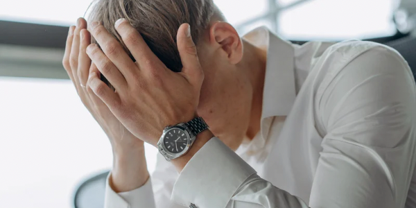 A person in business attire sits at a desk with their head in their hands, clearly overwhelmed or anxious. The image captures the physical expression of emotional and mental stress—tightened posture, closed-off body language, and visible fatigue—representing the unseen toll fear and chronic stress take on the body.