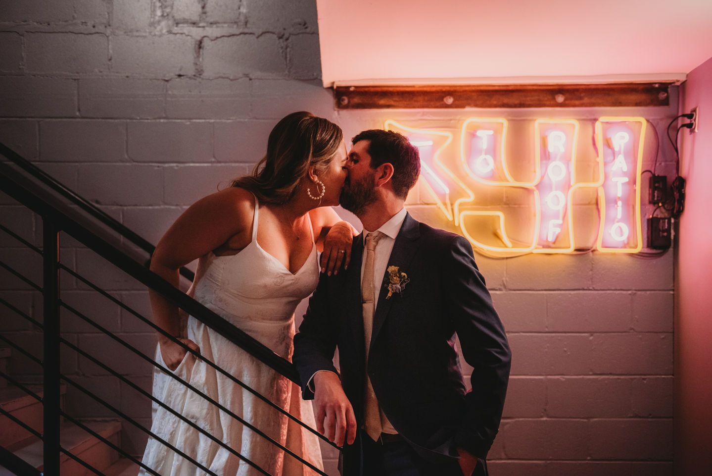the bride and groom kissing in a stairwell with a white concrete wall and a neon sign in the background