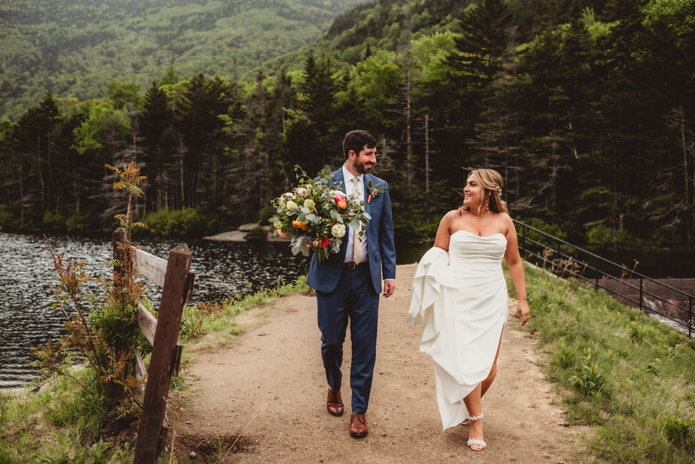 a bride and groom holding flowers walking past a stream on a dirt trail