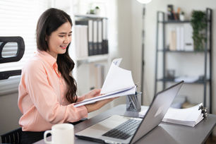 Eine junge Frau in einem Büro vor einem Laptop, welche Akten studiert. Sie lächelt und prüft eventuell ihre Post, welche sie auch digital erhalten hat. Service für KMU's etc.