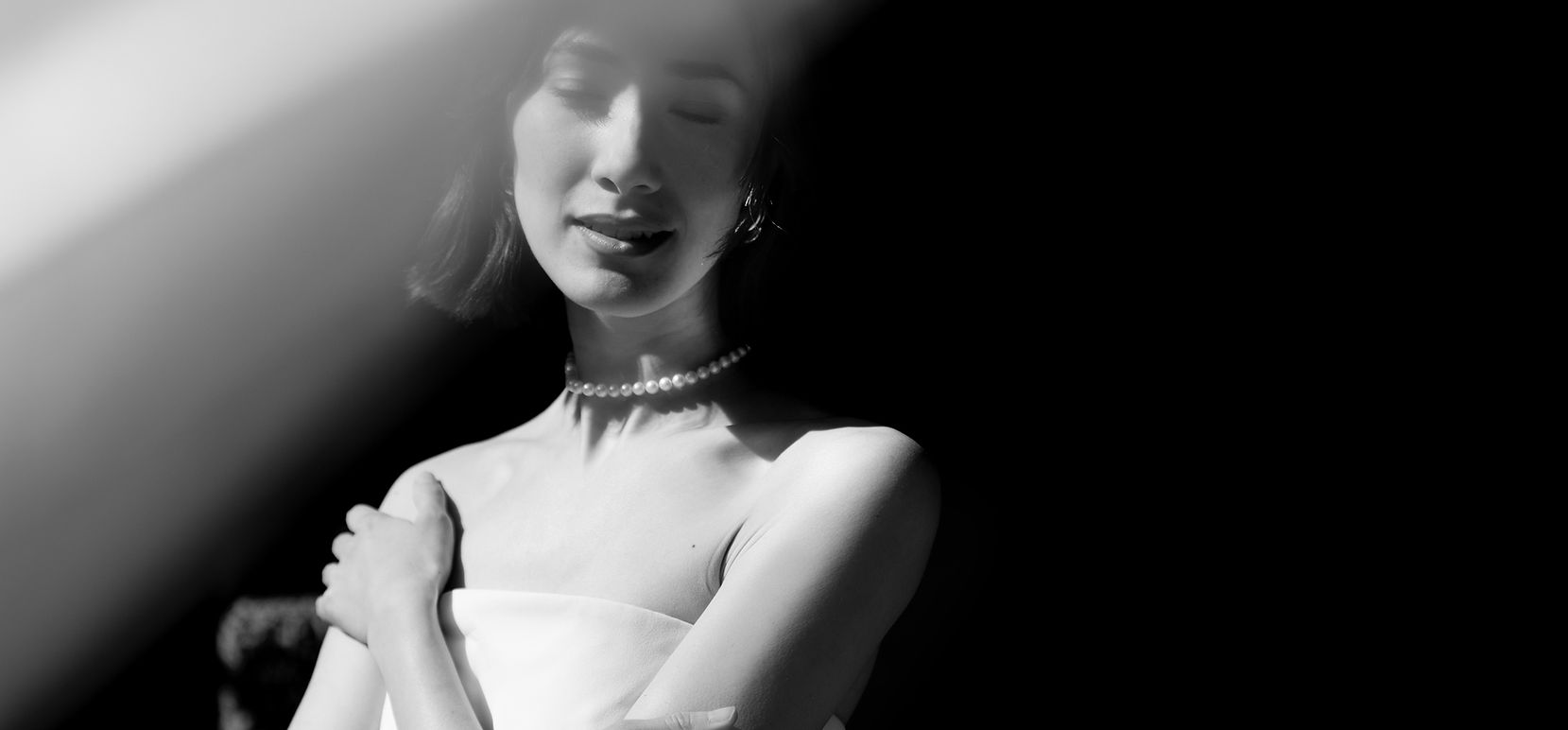 Black-and-white close-up portrait of a bride wearing a pearl necklace and a strapless top, softly illuminated by dramatic light and shadow with eyes gently closed.