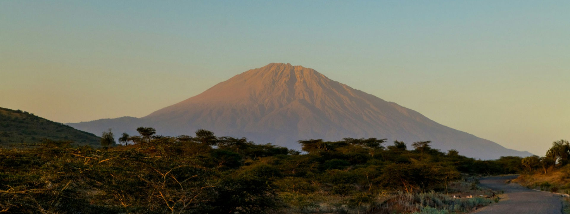 A slideshow image, featuring a distant view of Mount Meru rising behind a tree-lined foreground, during a sunrise.