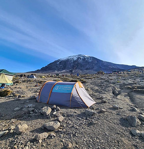 A thumbnail image, featuring a campsite, with a single tent in the foreground, displaying a green flag featuring the yellow Stunning Kilimanjaro logo, standing out against the rugged landscape.