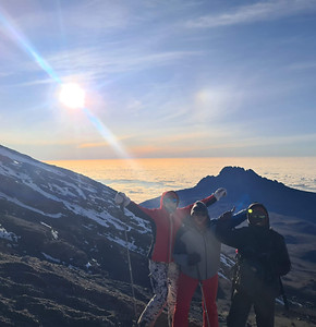 A thumbnail image, featuring three hikers standing on top of Mount Kilimanjaro, with in the background the view of a sea of clouds beneath a blue sky with a shining sun.
