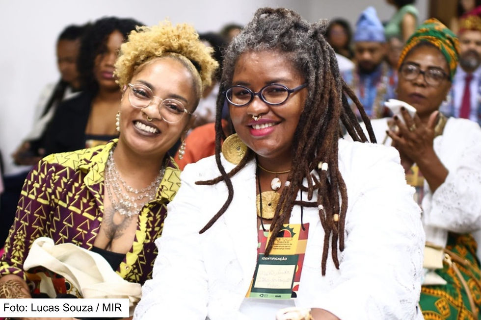 Foto de duas mulheres negras sorrindo, vestidas com trajes sociais, sentadas em uma plenária com diversas outras pessoas negras também sentadas atrás delas.