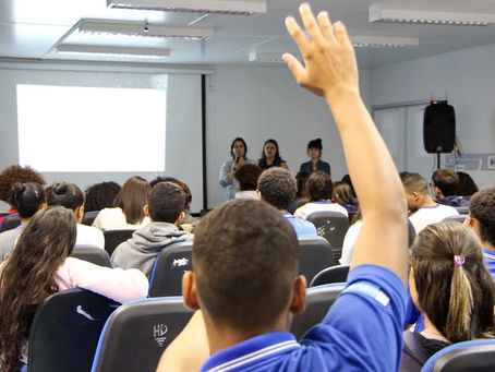 Foto a partir do fundo de uma sala mostra jovens sentados, sendo que um está com a mão levantada, como se estivesse pedindo para falar. Na frente, há mais três mulheres jovens, sendo que uma está falando em um microfone.