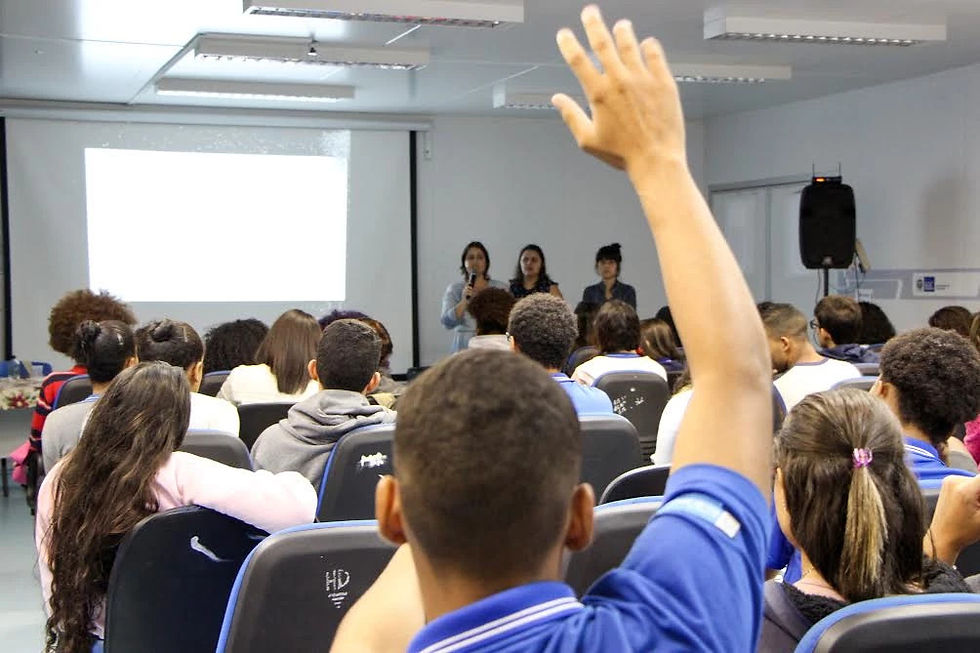 Foto a partir do fundo de uma sala mostra jovens sentados, sendo que um está com a mão levantada, como se estivesse pedindo para falar. Na frente, há mais três mulheres jovens, sendo que uma está falando em um microfone.