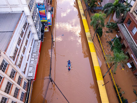 Foto de visa áerea de rua com enchente, estando visível os prédios ao lado. No centro da rua, cheia de uma água barrenta, há um caiaque com duas pessoas remando.