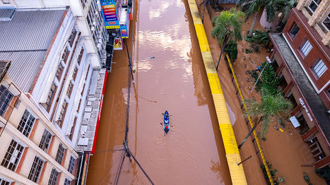 Foto de visa áerea de rua com enchente, estando visível os prédios ao lado. No centro da rua, cheia de uma água barrenta, há um caiaque com duas pessoas remando.