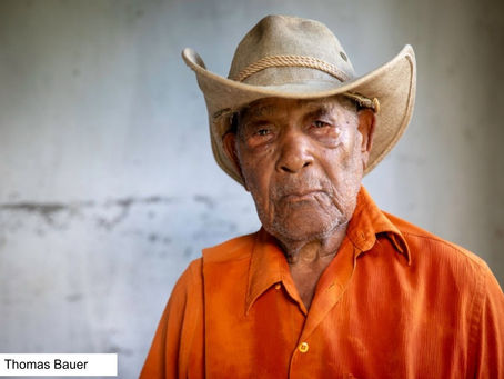 Foto do agricultor José Germano Lima, o Butinga, aos 91 anos. Ele está em frente a uma parede cinza, veste uma camisa laranja de botão e usa um chapéu de vaqueiro.