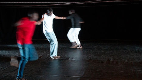 Foto de um espetáculo de dança, com três homens de mãos dadas em palco na cor preta e sob cordas estendidas à altura da cabeça.