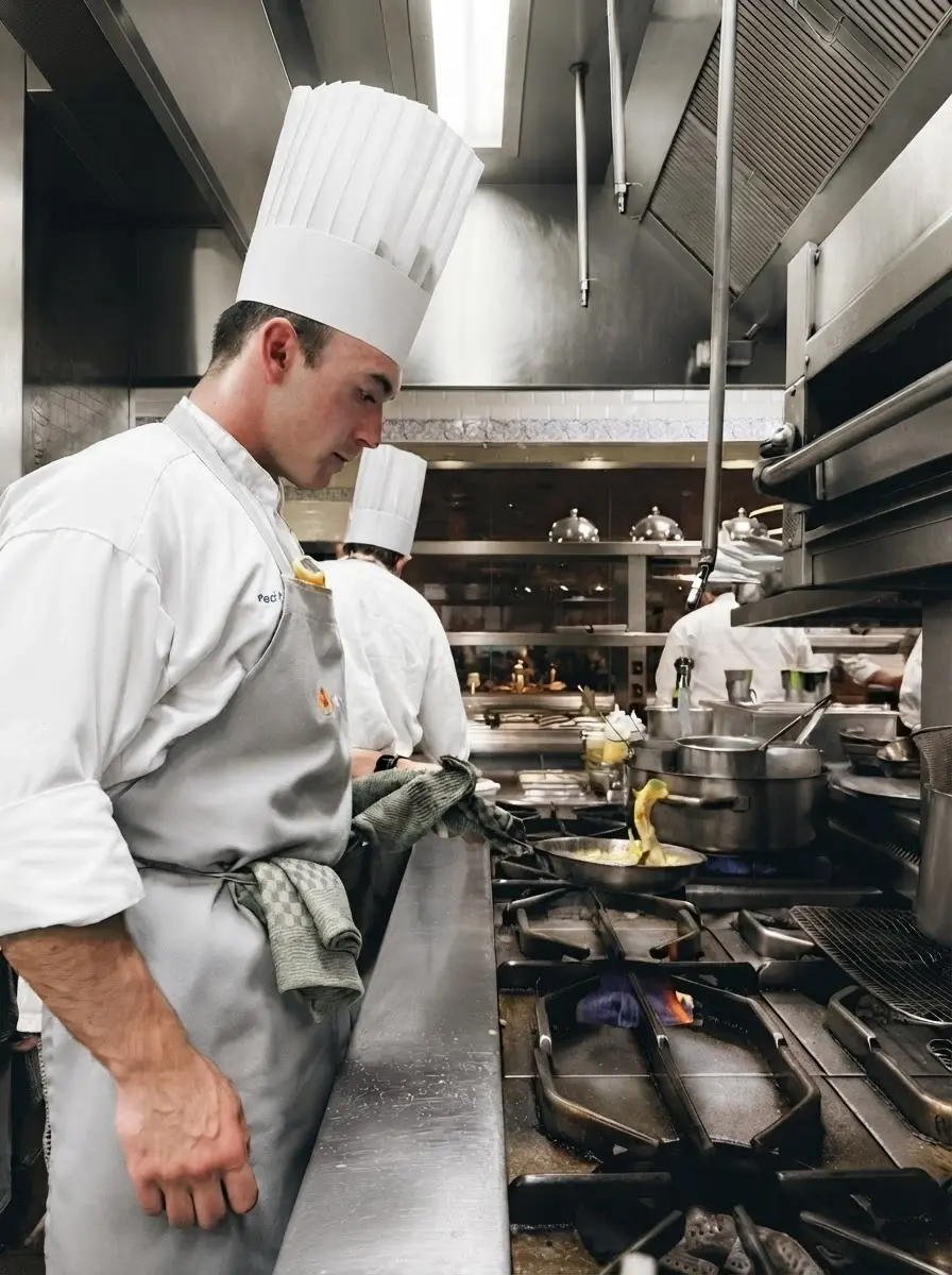 Studio photography of a focused chef wearing a white uniform cooking in a busy restaurant kitchen. The composition conveys a professional, high-energy atmosphere.